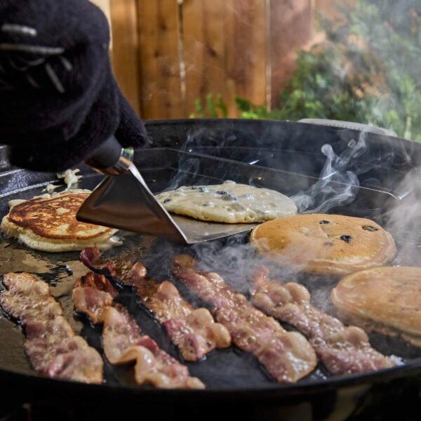 Mano con guante utilizando una espátula para voltear tortitas con arándanos en una plancha caliente mientras se cocinan tiras de tocino.