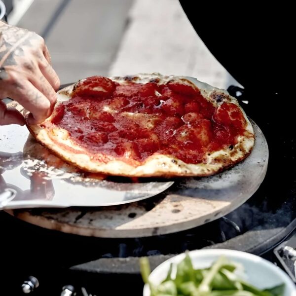 Pizza cocinándose sobre la piedra de pizza The Bastard en una parrilla, con base crujiente y salsa de tomate.