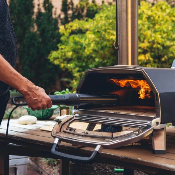 Persona utilizando el Looft Air Lighter 2 para encender un horno de pizza al aire libre, con llamas visibles en el interior del horno.