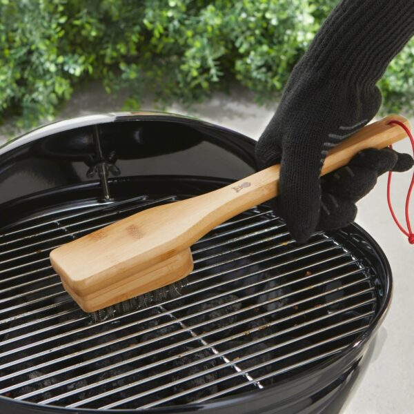 Persona con guantes de protección limpiando una parrilla con el cepillo de bambú Weber, eliminando residuos de la rejilla.