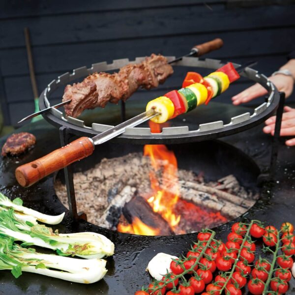 Brochetas de carne y vegetales asándose en anillo de brochetas OFYR sobre fuego de leña, con vegetales frescos en la parrilla.