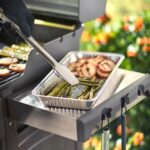 Espárragos y papas asadas en una bandeja de aluminio Weber junto a una parrilla encendida.