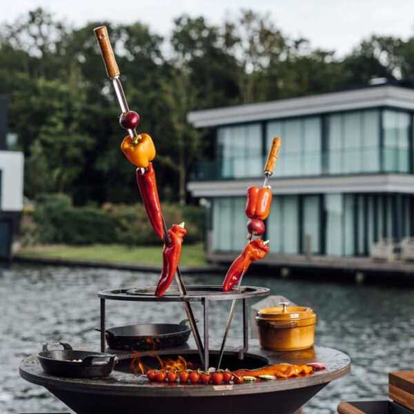 Brochetas de pimientos y cebollas asándose en parrilla OFYR junto a una cazuela y tomates cherry sobre fuego de leña.