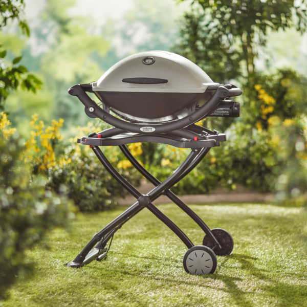 Barbacoa Weber Q1000/2000 en carrito plegable negro con ruedas, colocada en un jardín verde