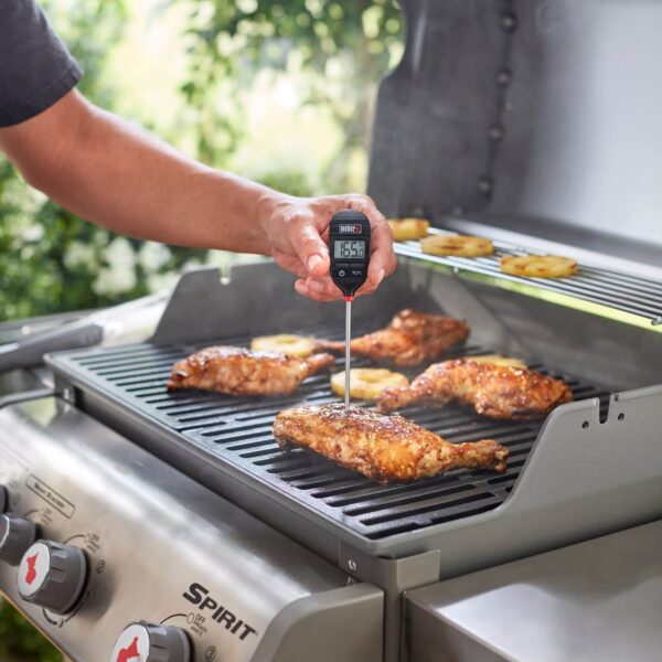 Mano sosteniendo un termómetro digital de lectura instantánea, midiendo la temperatura de un muslo de pollo a la parrilla en una barbacoa.