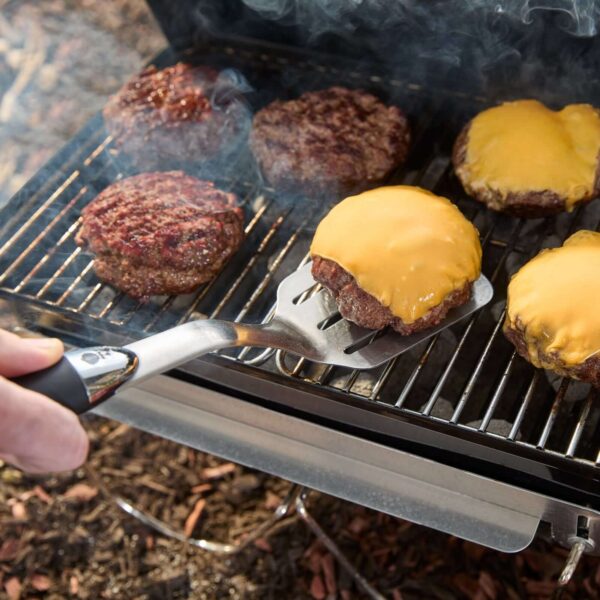 Espátula de precisión Weber levantando una hamburguesa con queso derretido en una parrilla, rodeada de otras hamburguesas en cocción con humo saliendo.