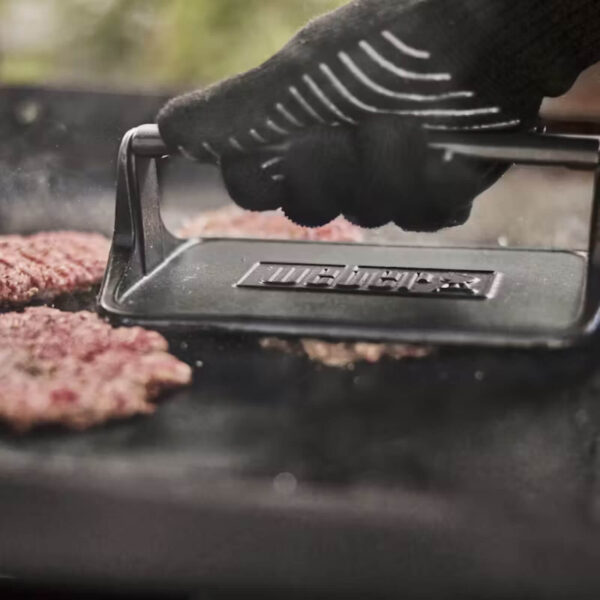 Primer plano de la prensa Weber aplastando una hamburguesa en una plancha caliente, con un guante térmico para mayor seguridad.