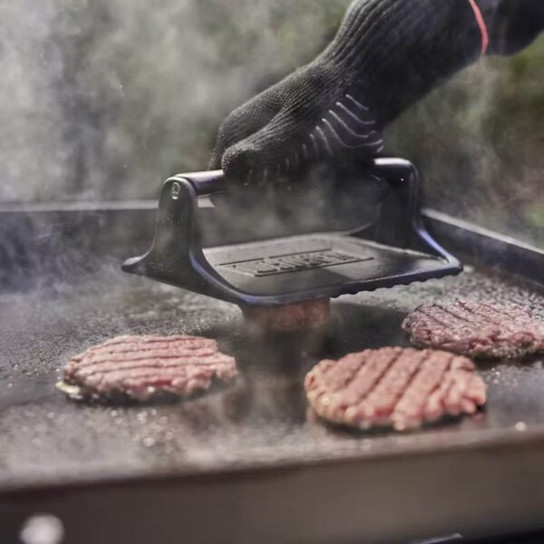 Prensa Weber utilizada para cocinar hamburguesas en una plancha caliente, con humo y un guante térmico para mayor seguridad.