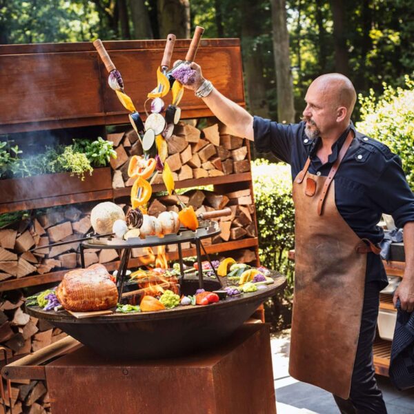 Hombre asando vegetales en una parrilla OFYR, usando un mandil de cuero marrón para protección.