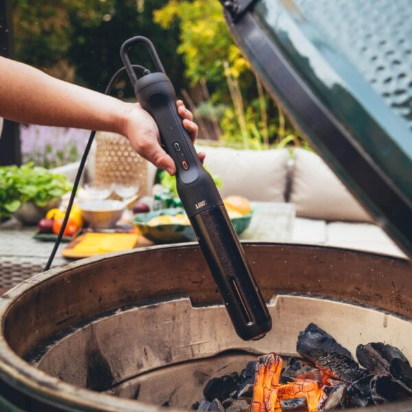 Mano utilizando el Looft Air Lighter 2 para encender carbón en una parrilla de cerámica al aire libre, con fondo de jardín y mesa con alimentos.