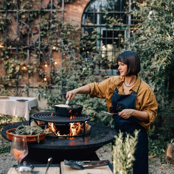 Mujer usando un mandil OFYR mientras cocina en una parrilla al aire libre, rodeada de vegetación.