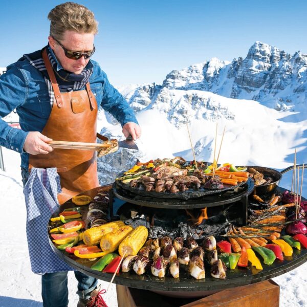 Hombre cocinando con parrilla OFYR en un entorno nevado, usando un mandil de cuero marrón para protección.