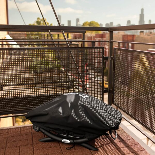 Barbacoa Weber® Q2000 cubierta con la funda Premium en un balcón urbano, con vista a la ciudad y protección contra el sol y el polvo.