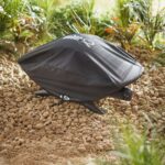 Funda Weber® Premium Q2000 cubriendo una barbacoa en un entorno natural con suelo de piedras y vegetación de fondo.
