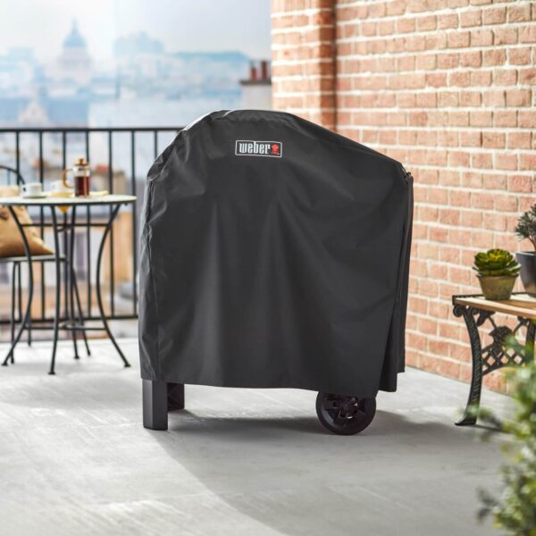 Funda Premium Pulse de Weber cubriendo una barbacoa con carrito en un balcón, con vista a la ciudad y decoración exterior.