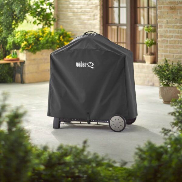 Funda Weber® Premium Q3000/Q2000 negra cubriendo una barbacoa con carro en un patio exterior, con logo blanco en la parte superior y ruedas visibles.