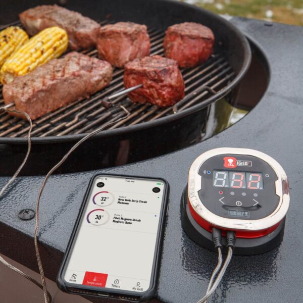 Termómetro iGrill 2 de Weber monitoreando la temperatura de varios cortes de carne en una parrilla, con lectura en un smartphone.