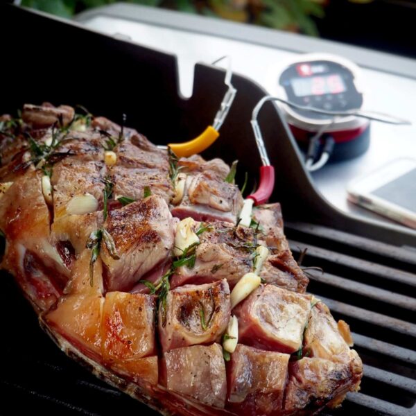 Corte de carne en la parrilla con sondas del iGrill 2 de Weber monitoreando la temperatura, con el termómetro y un smartphone en segundo plano.