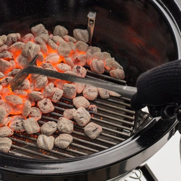 Persona con guante térmico usando una pala para carbón Weber para distribuir brasas encendidas en una parrilla de carbón.