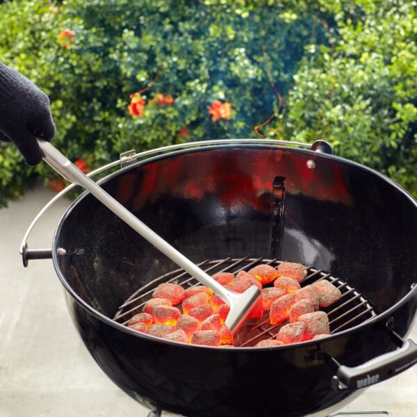 Persona con guante térmico usando una pala para carbón Weber en una parrilla de carbón encendida, distribuyendo las brasas incandescentes.