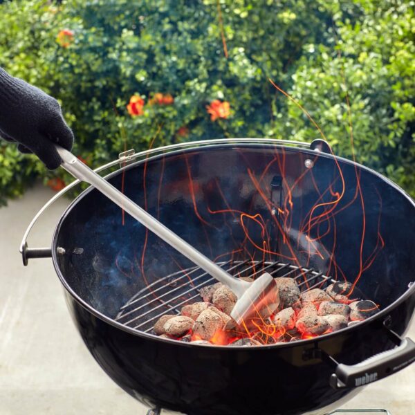 Persona con guante térmico usando una pala para carbón Weber en una parrilla de carbón encendida, con brasas incandescentes y chispas volando.