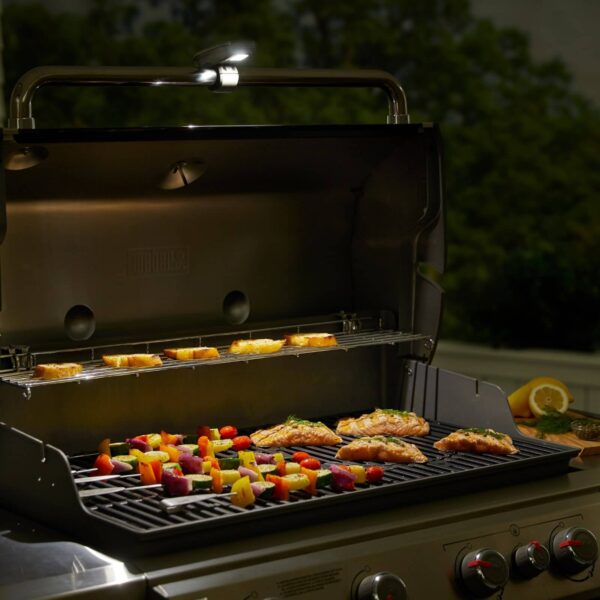 Lámpara Grill 'N Go de Weber iluminando una parrilla con salmón, brochetas de verduras y pan tostado durante la noche.