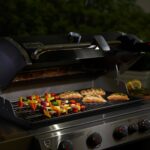 Lámpara Grill 'N Go de Weber iluminando una parrilla con salmón y brochetas de verduras durante la noche.