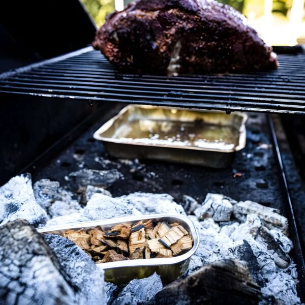 Bandeja Cook in Wood con astillas de madera colocada sobre brasas en una parrilla, con un corte de carne cocinándose en la rejilla superior.