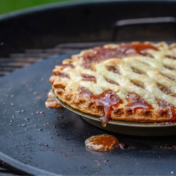 Tarta casera horneada sobre la piedra de barbacoa Weber, con masa dorada y relleno burbujeante derramándose.