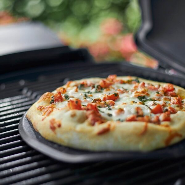 Pizza recién horneada sobre la piedra de barbacoa Weber en una parrilla, con fondo desenfocado de jardín.