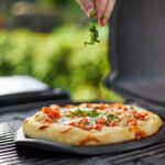 Mano agregando albahaca fresca sobre una pizza cocinada en la piedra de barbacoa Weber, en una parrilla al aire libre.