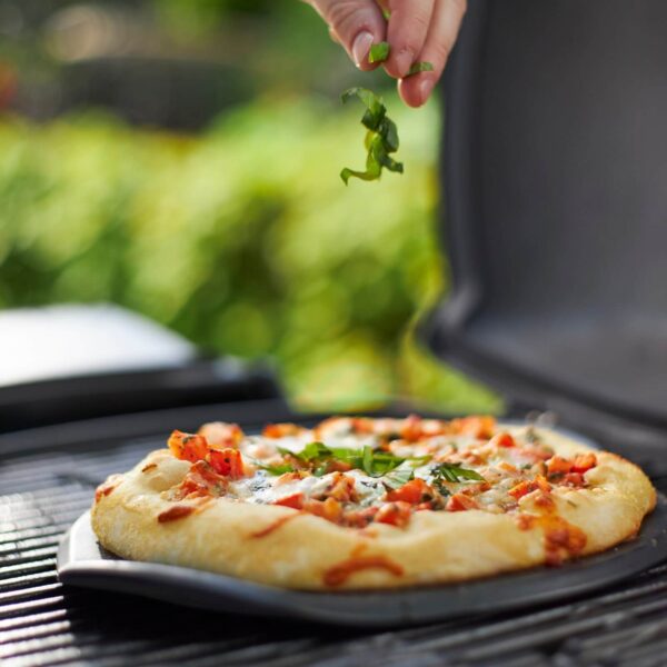 Mano agregando albahaca fresca sobre una pizza cocinada en la piedra de barbacoa Weber, en una parrilla al aire libre.