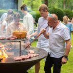 Grupo de personas disfrutando de una barbacoa al aire libre, mientras un hombre usa el Juego Buffadoo para avivar el fuego.