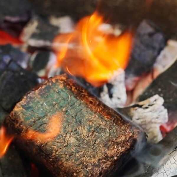 Bloque de madera comprimida ardiendo sobre brasas en una parrilla, liberando humo para el ahumado de alimentos.