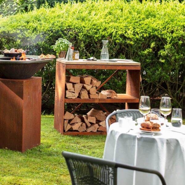 Almacén de Leña Dressoir de acero corten con tabla de teca en un jardín, junto a una barbacoa.