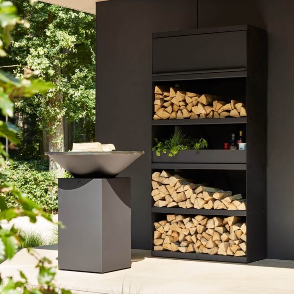 Almacén de Leña OFYR Black en un entorno exterior con parrilla OFYR, leña apilada y plantas decorativas, ideal para espacios modernos.