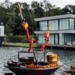 Brochetas de pimientos y cebollas rojas asándose en una parrilla Ofyr al aire libre, con vistas a un lago y una moderna casa de fondo.
