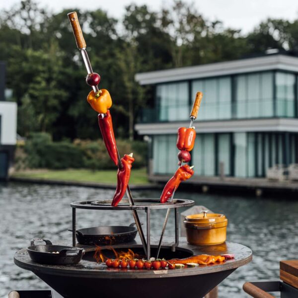 Brochetas de pimientos y cebollas rojas asándose en una parrilla Ofyr al aire libre, con vistas a un lago y una moderna casa de fondo.