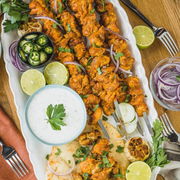 Brochetas de acero inoxidable con pollo especiado, servidas en una bandeja con salsa blanca, rodajas de limón, pan naan y vegetales frescos.