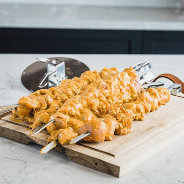 Brochetas de acero inoxidable con trozos de pollo marinado en especias, colocadas sobre una tabla de madera, listas para asar.