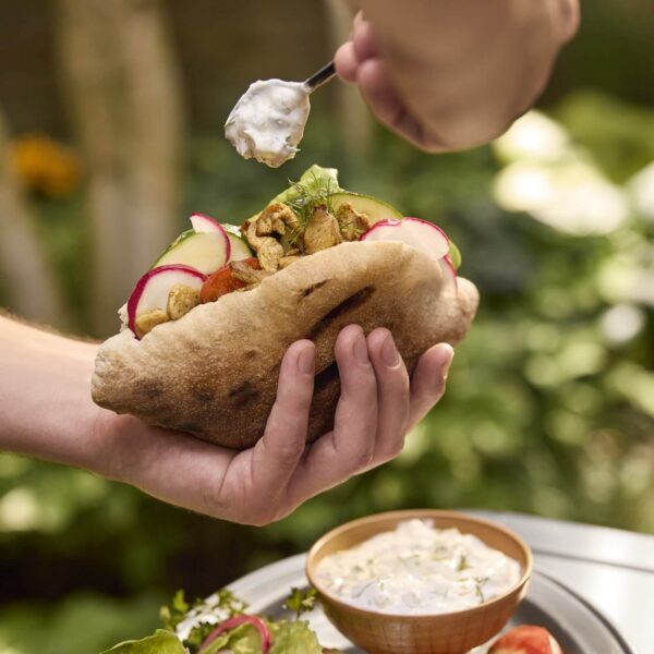 Pan pita relleno de vegetales frescos, pollo asado y salsa blanca siendo añadida con una cuchara.