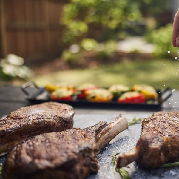 Costillas de res asadas con sal gruesa, presentadas en un entorno al aire libre con pimientos rellenos al fondo.