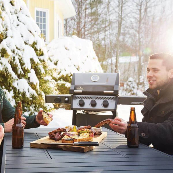 Tres amigos compartiendo una comida junto a una parrilla en un entorno nevado y soleado, con bebidas y una tabla de alimentos en la mesa.