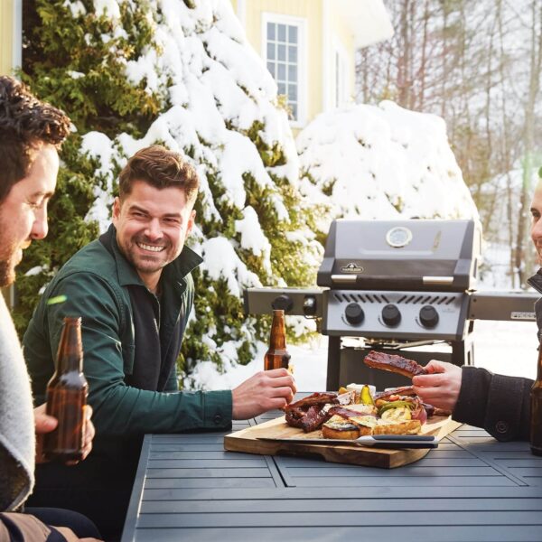 Grupo de amigos disfrutando de comida y bebidas junto a una parrilla en un ambiente al aire libre con nieve de fondo.