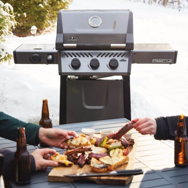 Parrilla de gas en un entorno nevado con una mesa al frente donde se comparte una tabla de alimentos a la parrilla y botellas de cerveza.