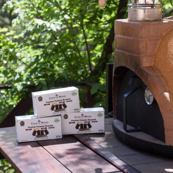 Tres cajas de Foodie Power Blocks de Cook in Wood colocadas sobre una mesa de madera junto a un horno de exterior, en un entorno natural con árboles al fondo.