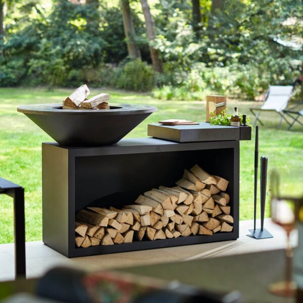 Barbacoa de leña Isla OFYR en acero negro con encimera de cerámica gris oscuro y compartimento para leña en un jardín exterior.