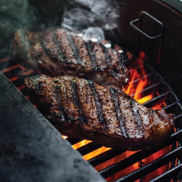 Dos filetes cocinándose en una parrilla Kamado Joe Classic Joe, con llamas y marcas de asado visibles.