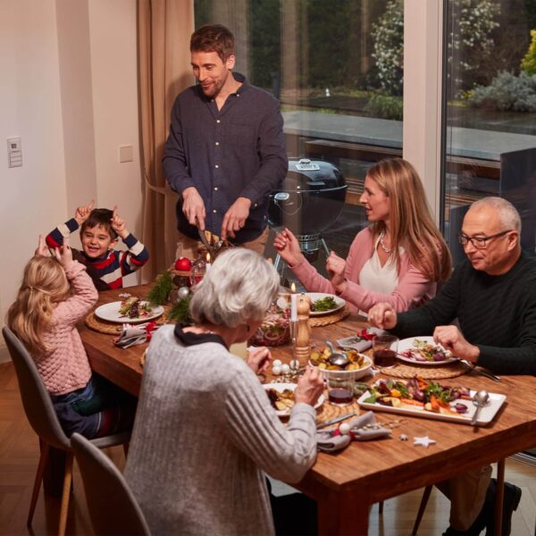 Cena familiar con niños jugando en la mesa y la barbacoa Weber Master-Touch Premium visible en el exterior.