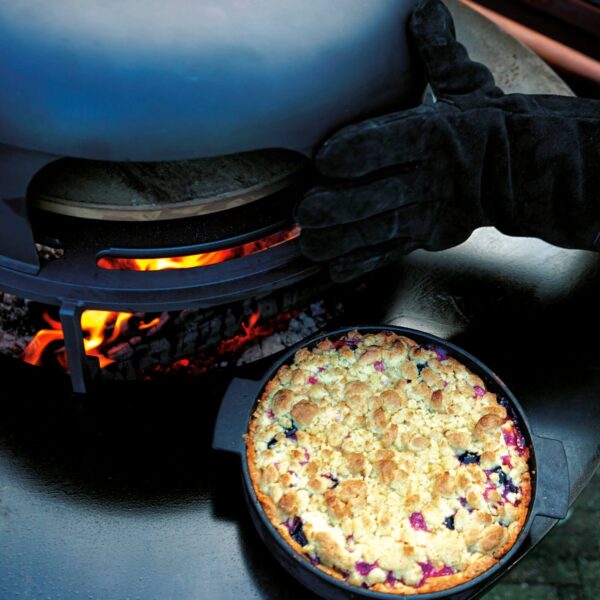 Pastel crumble de frutas cocinándose en el horno de pizza OFYR, con guantes resistentes al calor ajustando el cloche, mostrando la versatilidad para recetas dulces.